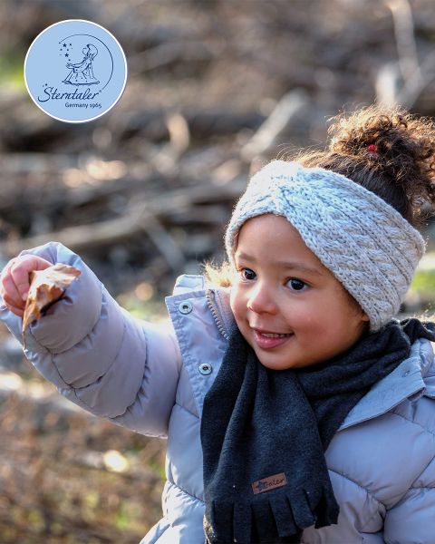Una niña pequeña con un abrigo gris claro, una bufanda gris y una diadema de punto sostiene una hoja afuera. El fondo está borroso por ramas y luz solar. En la esquina se ve un logo redondo que dice "Sterntaler Alemania 1965".