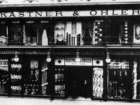 Schwarz-Weiß-Foto einer alten Ladenfront mit dem Schild „Kastner & Ohler“. Die Fassade zeigt in den Fenstern verschiedene Stoffe und Textilien, über dem Eingang hängen mehrere Hängelampen.