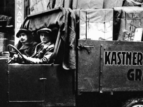Zwei Personen sitzen in einem mit großen Paketen beladenen Oldtimer-LKW. Auf der Seite des LKWs ist der teilweise sichtbare Text „KASTNER“ zu sehen. Die Szene scheint historisch zu sein, da beide Personen Mützen und Mäntel tragen.