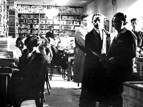 Ein Schwarzweißfoto von Menschen, die in einem Archivraum voller Regale mit Dokumenten und Kartons arbeiten. Einige Personen sitzen an Tischen und sortieren Papiere, andere stehen. Die Atmosphäre wirkt geschäftig und konzentriert.