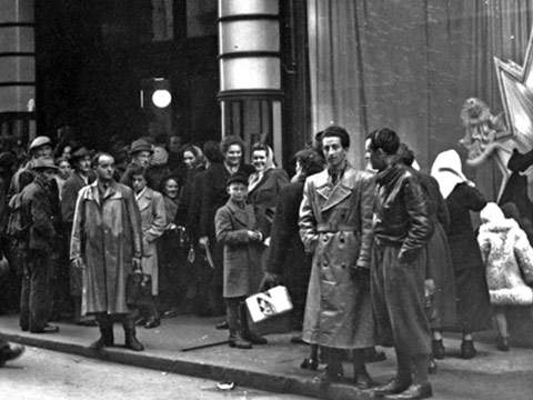 Ein Schwarzweißfoto einer Menschenmenge, die sich vor einem Gebäude versammelt hat. Die Menschen tragen Kleidung aus der Mitte des 20. Jahrhunderts, darunter Mäntel und Hüte. Einige schauen in die Kamera, während andere miteinander reden. Im Hintergrund ist ein großes Schaufenster zu sehen.