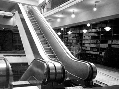 Schwarzweißfoto des Interieurs eines alten Kaufhauses mit einem Rolltreppendesign aus der frühen Zeit. Im Hintergrund sind Regale voller Kisten zu sehen und von der Decke hängen Lampen.