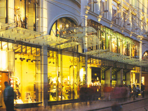 Eine belebte Stadtstraße bei Nacht mit verschwommenen Fußgängern, die an einem hell erleuchteten, stilvollen Laden vorbeigehen. Große Fenster zeigen verschiedene Schaufensterpuppen und Waren, während dekorative Lichter die Fassade des Gebäudes beleuchten.