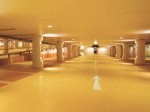 Eine hell erleuchtete Tiefgarage mit gelben Wänden und Boden. Weiße Pfeile auf dem Boden leiten den Verkehr und mehrere Säulen stützen die Decke. Der Raum ist leer und bietet einen klaren Blick auf das weitläufige Gelände.