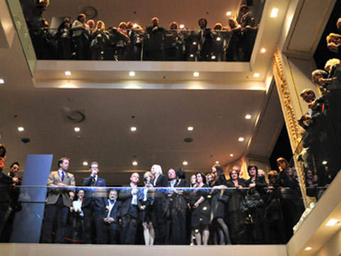 Eine große Gruppe von Menschen in formeller Kleidung steht auf zwei Ebenen eines hell erleuchteten Innenbalkons und blickt nach unten. Die Atmosphäre ist lebhaft, die Menschen unterhalten sich und beobachten etwas unten.