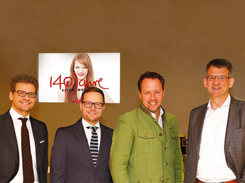 Vier Männer in Abendgarderobe stehen lächelnd vor einer braunen Wand. Hinter ihnen hängt ein Plakat mit einer Frau mit roten Haaren und dem Text „140 Jahre Alles Mode“.