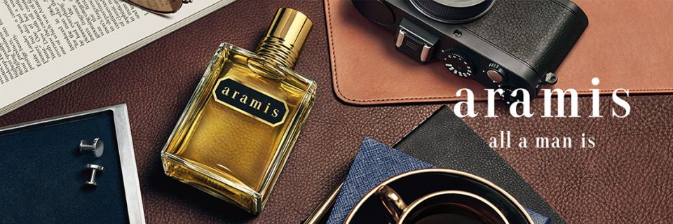 Representación plana de una disposición elegante con una botella de perfume Aramis, una cámara, gemelos, un libro abierto, un cuaderno y una taza de café sobre una superficie marrón texturizada. El texto dice: "Aramis es todo lo que un hombre es".