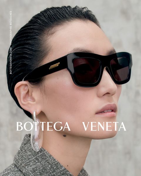 Persona con el pelo peinado hacia atrás, grandes gafas de sol negras y aros transparentes extragrandes. El texto dice "Bottega Veneta". La persona mira ligeramente hacia un lado frente a un fondo neutro.
