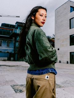 Una mujer con cabello largo y oscuro, una chaqueta acolchada verde y pantalones de color marrón claro está de pie al aire libre en un entorno urbano y mira por encima del hombro.