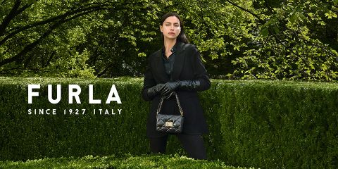 Una mujer está de pie frente a una exuberante vegetación. Lleva un atuendo negro y guantes, y sostiene un bolso negro acolchado en la mano. A la izquierda se puede ver el texto "Furla Desde 1927 Italia".