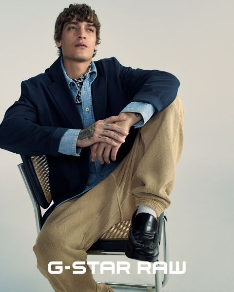 Una persona está sentada en una silla y lleva un blazer oscuro, una camisa vaquera y pantalones beige. Tiene tatuajes en la mano y mira hacia arriba. La persona también lleva zapatos bajos y en la parte inferior de la imagen aparece el texto "G-STAR RAW".