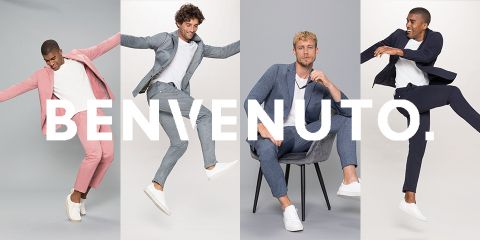 Se ven cuatro hombres en elegantes trajes y zapatillas blancas frente a un fondo brillante. Dos saltan enérgicamente, uno está tumbado en una silla y uno posa. La palabra "BENVENUTO" está superpuesta a la imagen.