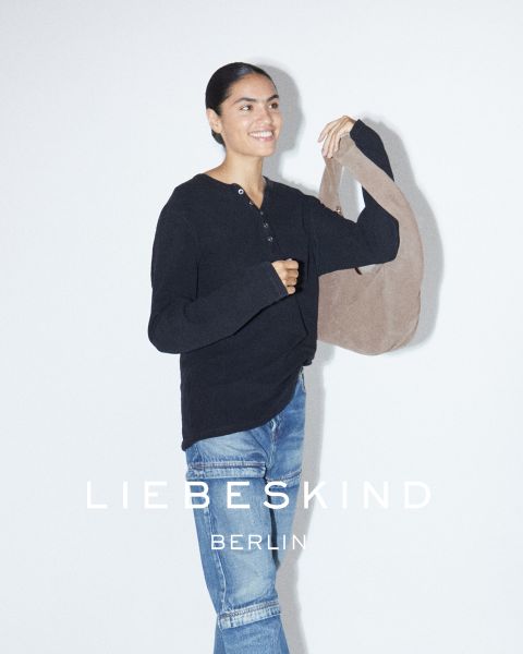 Una mujer sonriente con una camisa negra de manga larga y jeans lleva un bolso marrón claro sobre el hombro. El texto "Liebeskind Berlin" aparece en la parte inferior de la imagen.