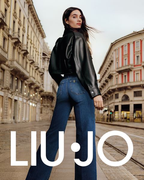 Una mujer está de pie en una calle de la ciudad. Lleva una chaqueta de cuero negra y vaqueros azules. Mira por encima del hombro mientras su largo cabello ondea al viento. En el fondo se puede ver arquitectura clásica. Abajo está el texto "LIU JO".