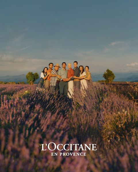 Un grupo de personas están juntas en un campo de lavanda, tomándose de las manos bajo un cielo despejado. Sonríen a la cámara, rodeados de brillantes flores púrpuras. Abajo se ve la marca "L'OCCITANE EN PROVENCE".