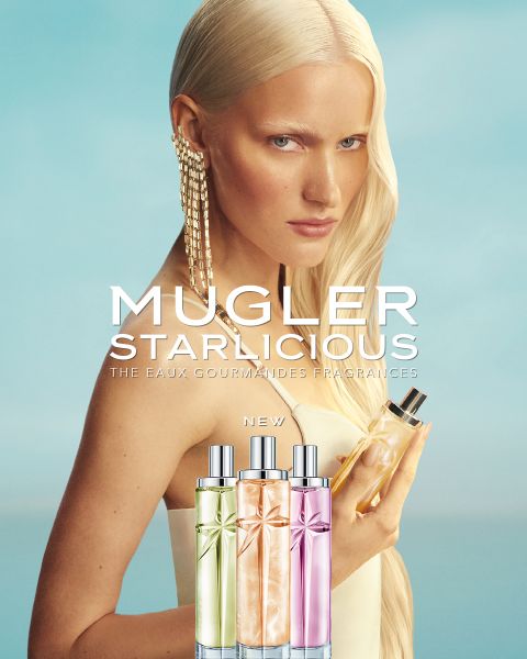 Una mujer con cabello largo y rubio sostiene un frasco de perfume en la mano. En la parte inferior de la imagen, se ven tres frascos coloridos de «Mugler Starlcious» frente a un cielo azul.