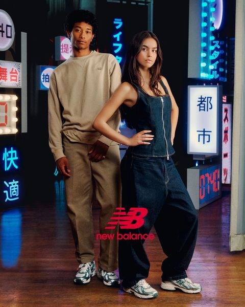Un hombre y una mujer posan frente a letreros de neón. El hombre lleva un chándal beige y la mujer un mono vaquero. Ambos llevan zapatillas New Balance. La escena está iluminada por varias luces de neón en azul y rojo.