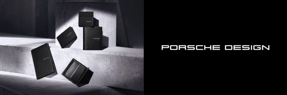 Una colección de accesorios de cuero negro de Porsche Design, que incluye billeteras y tarjeteros, se presenta elegantemente sobre superficies de hormigón, proyectando suaves sombras. El logotipo de Porsche Design se muestra prominentemente en el lado derecho.