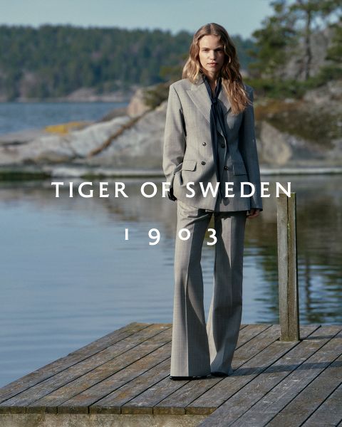 Una mujer con un traje gris está de pie en un muelle de madera en un lago con orillas rocosas y arboladas. El texto "TIGER OF SWEDEN 1903" está escrito a través de la imagen.