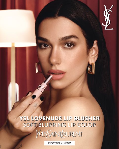 Una mujer aplicándose lápiz labial rosa y llevando aretes de aro dorados, frente a cortinas rojas. El texto promociona el «YSL Lovenuede Lip Blusher», un lápiz labial de difuminado suave.