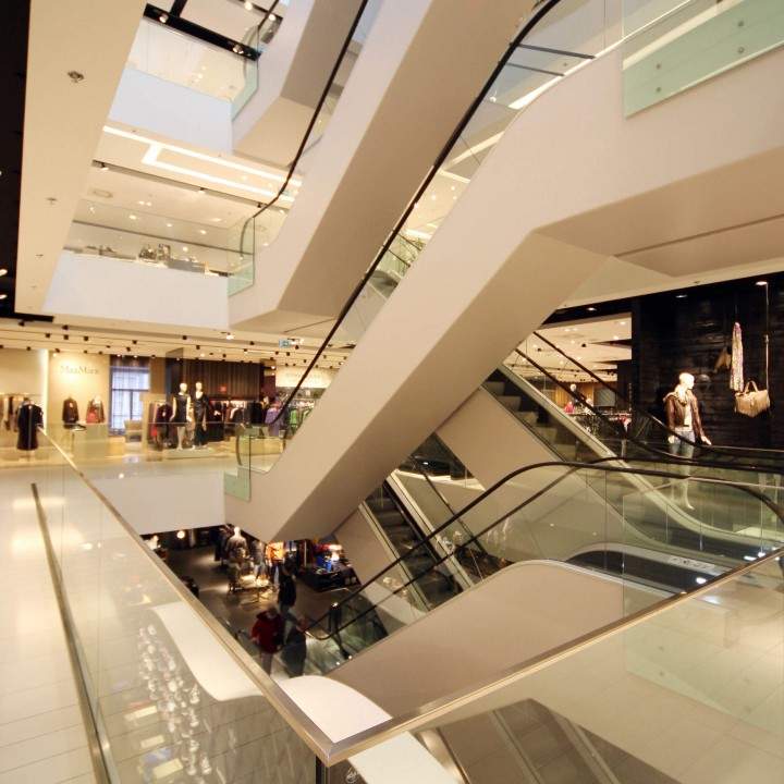 Un moderno centro comercial con múltiples pisos y escaleras mecánicas. El espacio está bien iluminado y cuenta con tiendas con escaparates en diferentes niveles. Se pueden observar barandillas de vidrio y elegantes elementos de diseño arquitectónico en todas partes.