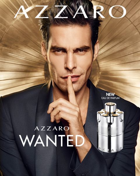 Un hombre en traje se lleva un dedo a los labios en un gesto tranquilizador. Delante de él hay una botella de Azzaro Wanted Eau de Parfum. El fondo muestra un diseño metálico circular. El texto dice "Azzaro" y "Wanted".