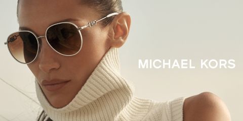Una persona con gafas de sol redondas y un jersey de cuello alto blanco mira hacia un lado. El fondo está suavemente borroso y a la derecha se ve el nombre "Michael Kors" en letras blancas.