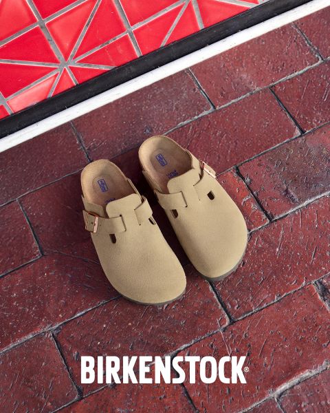 Un par de zuecos Birkenstock color beige con correas de hebilla sobre un suelo de ladrillo rojo junto a una pared de azulejos rojos con diseño geométrico. El logotipo blanco de Birkenstock está en la parte inferior de la imagen.