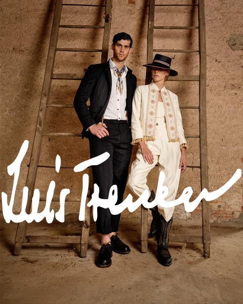 Un hombre y una mujer con elegantes atuendos de inspiración vintage están de pie entre dos escaleras de madera frente a una pared rústica. El hombre lleva un traje de rayas oscuras, la mujer un traje color crema con sombrero y botas. "Luis Trenker" está escrito en negrita.