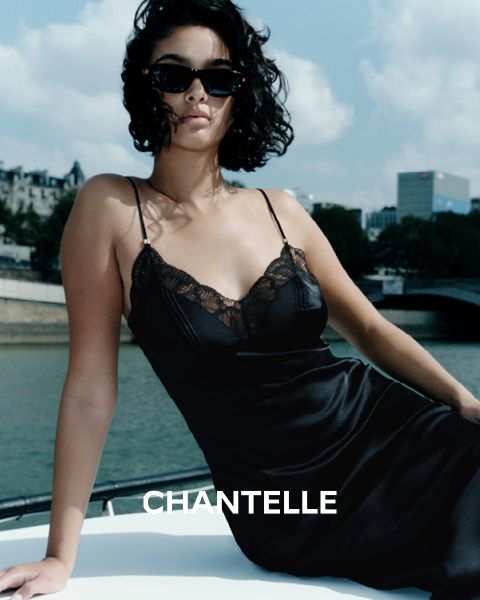 Una mujer con un vestido lencero negro de encaje y gafas de sol sentada en un barco cerca de un río urbano, con edificios y árboles al fondo. La palabra "CHANTELLE" se puede leer en la parte inferior de la imagen.