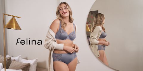 Una mujer en ropa interior gris está de pie sonriendo en una habitación con un espejo redondo. Lleva una chaqueta de punto beige y mira hacia otro lado del espejo. A su lado hay una lámpara de pie moderna y dorada. A la izquierda se puede ver la palabra "Felina".