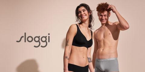 Una mujer sonriente y un hombre en ropa interior de Sloggi están de pie frente a un fondo neutro con el logo de la marca en el lado izquierdo. La mujer lleva un conjunto negro y el hombre un slip gris.