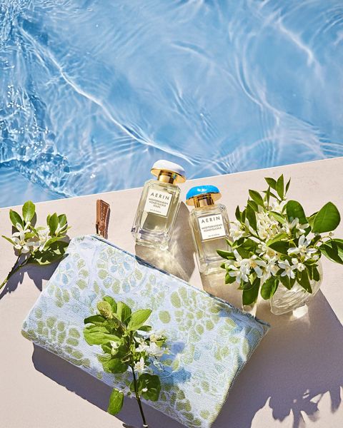 Dos frascos de perfume, una bolsa decorada con flores y pequeños jarrones con flores blancas están dispuestos en una repisa al borde de la piscina, cerca del agua azul clara.