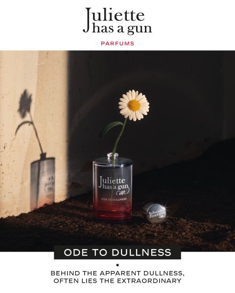 En una repisa hay un frasco de perfume de vidrio transparente con las inscripciones "Juliette has a gun" y "ODE TO DULLNESS". Dentro del frasco hay una sola margarita blanca. La sombra de la margarita se proyecta sobre una pared detrás. La luz del sol crea un efecto dramático.