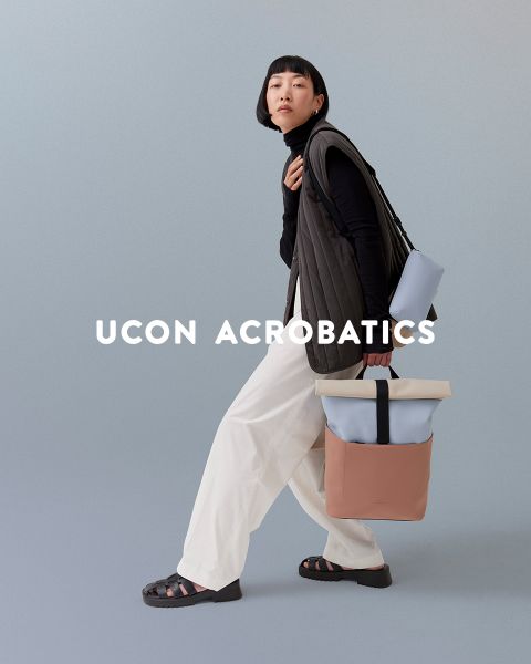 Una persona con corte de pelo bob posa frente a un fondo cubierto. Lleva un top negro, pantalones blancos y sandalias, y lleva una mochila elegante. Encima de la imagen pone "Ucon Acrobatics".