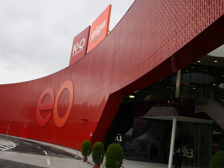 Un edificio grande y moderno con una fachada roja y ondulada y las letras distintivas "eo". La entrada tiene puertas de cristal y plantas en macetas. En el fondo se ve un cielo nublado.