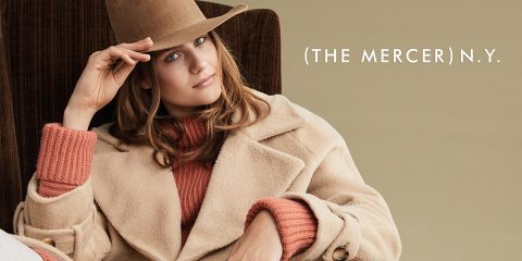Una persona con un abrigo y un sombrero beige está sentada en un sillón marrón. A su lado está el texto "The Mercer N.Y." sobre un fondo beige claro.