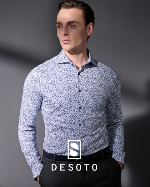 Un hombre con una camisa estampada de manga larga y pantalones oscuros está de pie frente a un fondo gris. Mira hacia un lado con las manos en los bolsillos. La inscripción "DESOTO" y el logotipo de DESOTO se ven en la parte inferior.
