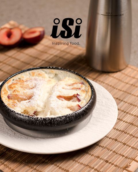 Un postre horneado espolvoreado con azúcar glas se sirve en un cuenco negro, al fondo se ven ciruelas y un dispensador de metal.