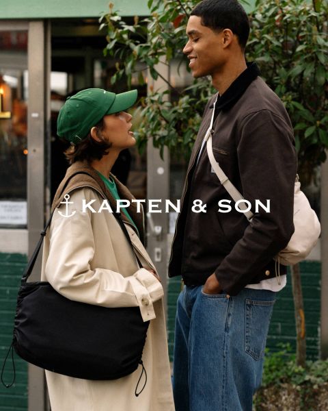Una mujer y un hombre se sonríen al aire libre. La mujer lleva un gorro verde y un abrigo beige y sostiene un bolso negro en la mano. El hombre lleva una chaqueta marrón y un bolso color crema. En el centro se lee "Kapten & Son".