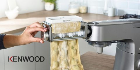 Una persona usa una máquina de cocina Kenwood con un accesorio para pasta para hacer ravioles. La masa fresca para pasta se extiende y se corta en trozos de ravioles. La cocina está modernamente amueblada, con vasos y una taza de café al fondo.