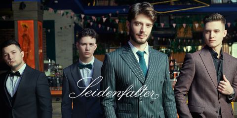 Cuatro hombres en trajes formales posan con confianza en un ambiente elegante con decoración colgante. A lo ancho de la imagen se lee la inscripción "Seidenfalter".