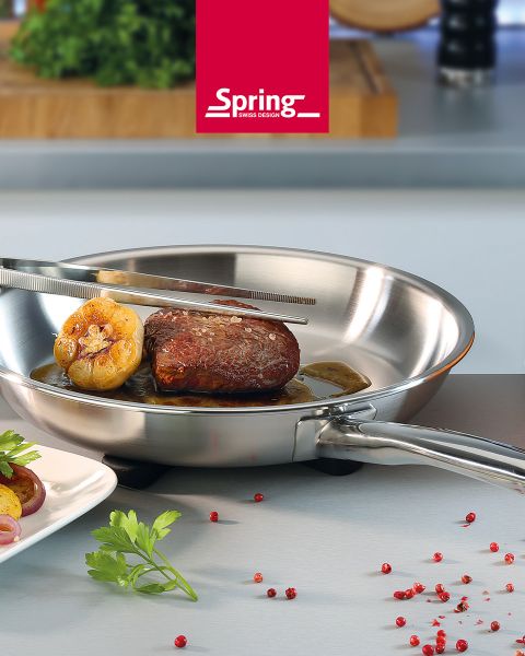 Sobre una encimera de cocina hay una sartén de acero inoxidable con un bistec y ajo, que está siendo volteado con unas pinzas. Al lado hay granos de pimienta roja y perejil.