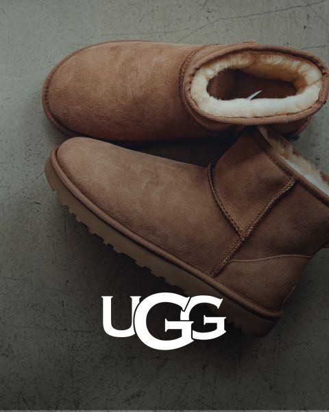 Un par de botas UGG de gamuza marrón con un forro interior de felpa, descansando sobre una superficie gris. El logo de UGG está prominentemente colocado en la parte inferior de la imagen.
