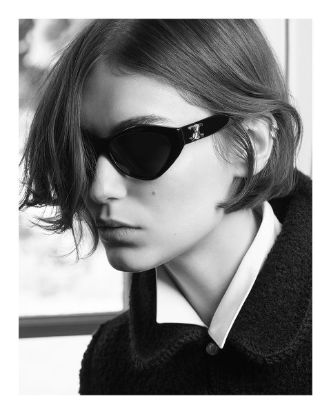 Fotografía en blanco y negro de una persona con cabello corto, gafas de sol oscuras y una camisa con cuello debajo de una chaqueta texturizada, mirando hacia un lado en un entorno con poca luz.