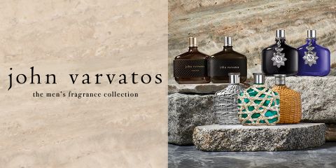 Una selección de seis fragancias masculinas de John Varvatos se exhibe sobre losas de piedra. Los frascos varían en diseño y color, desde patrones tejidos hasta superficies lisas y metálicas. El texto a la izquierda dice "John Varvatos, la colección de fragancias masculinas".