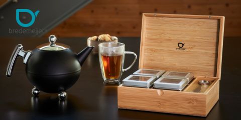 Sobre una mesa oscura hay una tetera negra, un vaso de cristal lleno de té y una caja de madera con cuatro latas de té. En la tetera y en la caja se puede ver el logo "Bredemeijer".
