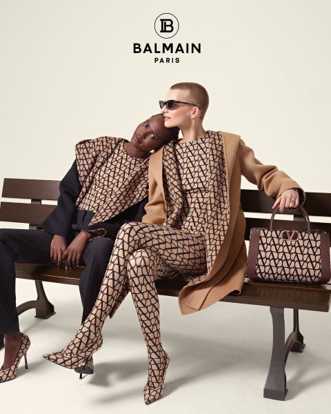 Dos modelos están sentadas en un banco con conjuntos Balmain a juego con estampados geométricos. Una se apoya en la otra, vestida con un top y pantalones negros, mientras que la otra lleva un abrigo color camel y gafas de sol. A su lado hay un bolso estampado. Arriba, el logotipo de Balmain.