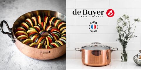 Una olla de cobre llena de rodajas de calabacín, berenjena y tomate en capas. Al lado hay una cacerola de cobre con tapa delante de una pared alicatada blanca con flores en un jarrón. El logotipo de de Buyer y el texto "Made in France" son visibles.
