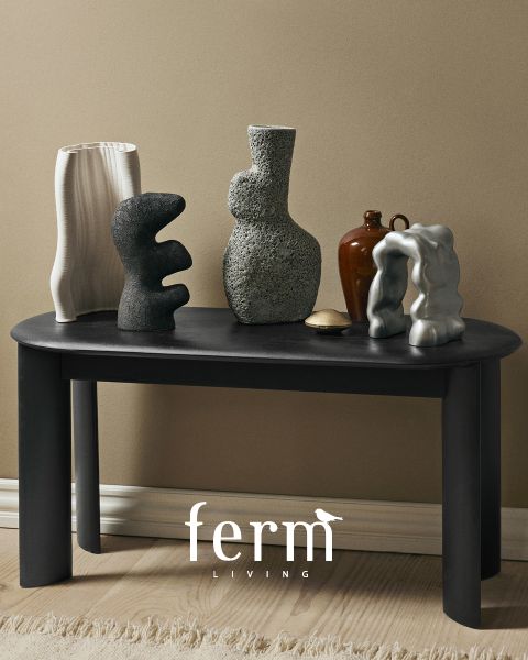 Una mesa negra con esculturas de cerámica abstractas en varias formas y texturas se encuentra frente a una pared beige. El suelo es de madera clara y en la parte inferior se ve el logo de "ferm LIVING".
